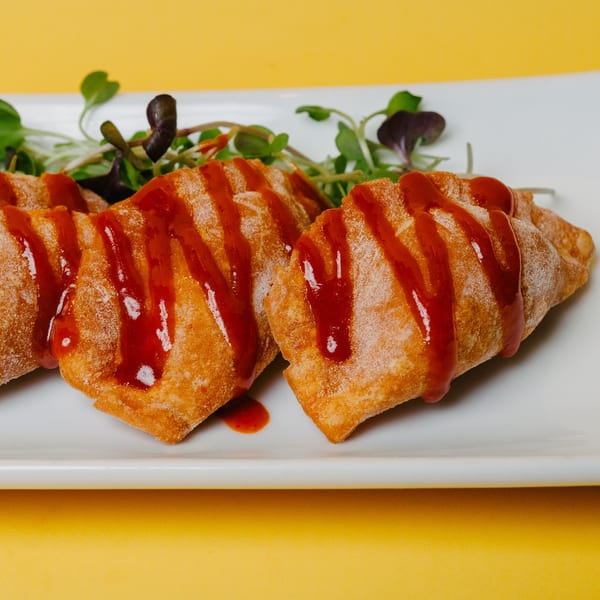 Three golden-fried dumplings drizzled with red sauce and garnished with microgreens on a white plate.