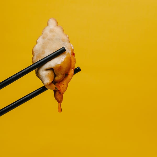 Chopsticks hold a fried dumpling dripping sauce against a vibrant yellow background.