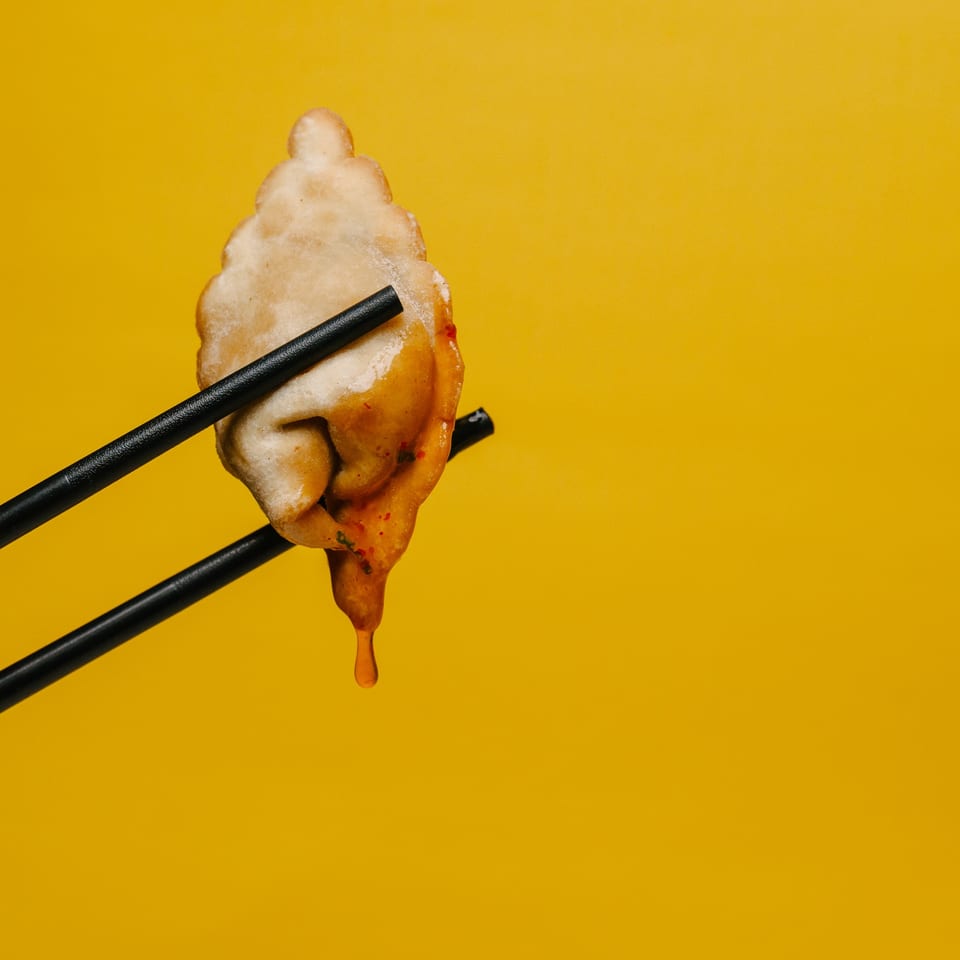 Chopsticks hold a fried dumpling dripping sauce against a vibrant yellow background.