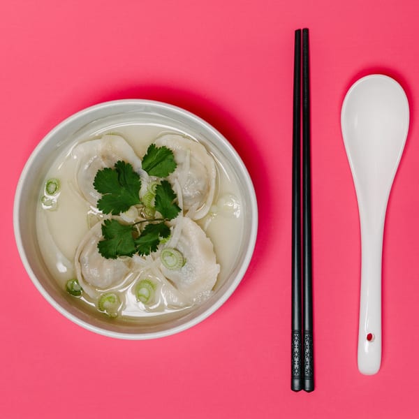 Noods To Go's dumpling soup with cilantro and scallions, chopsticks, and a spoon on a pink background.