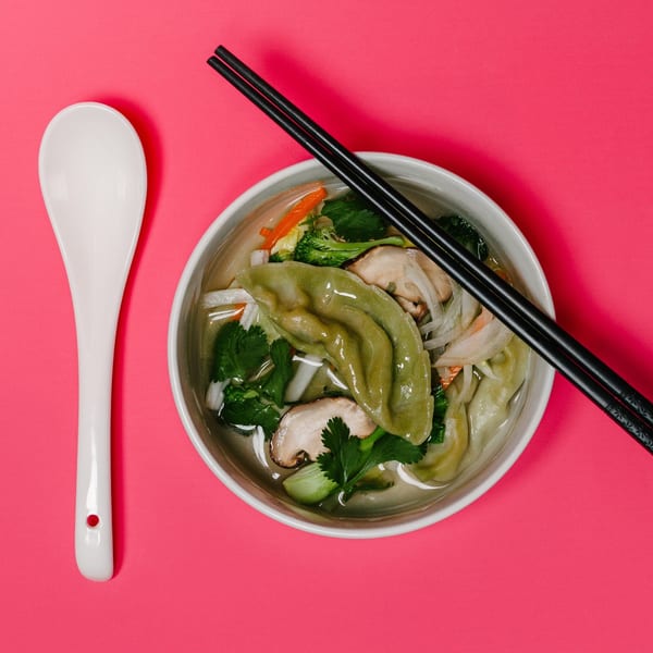 A bowl of Noods To Go dumpling soup with vegetables and chopsticks on a pink background.
