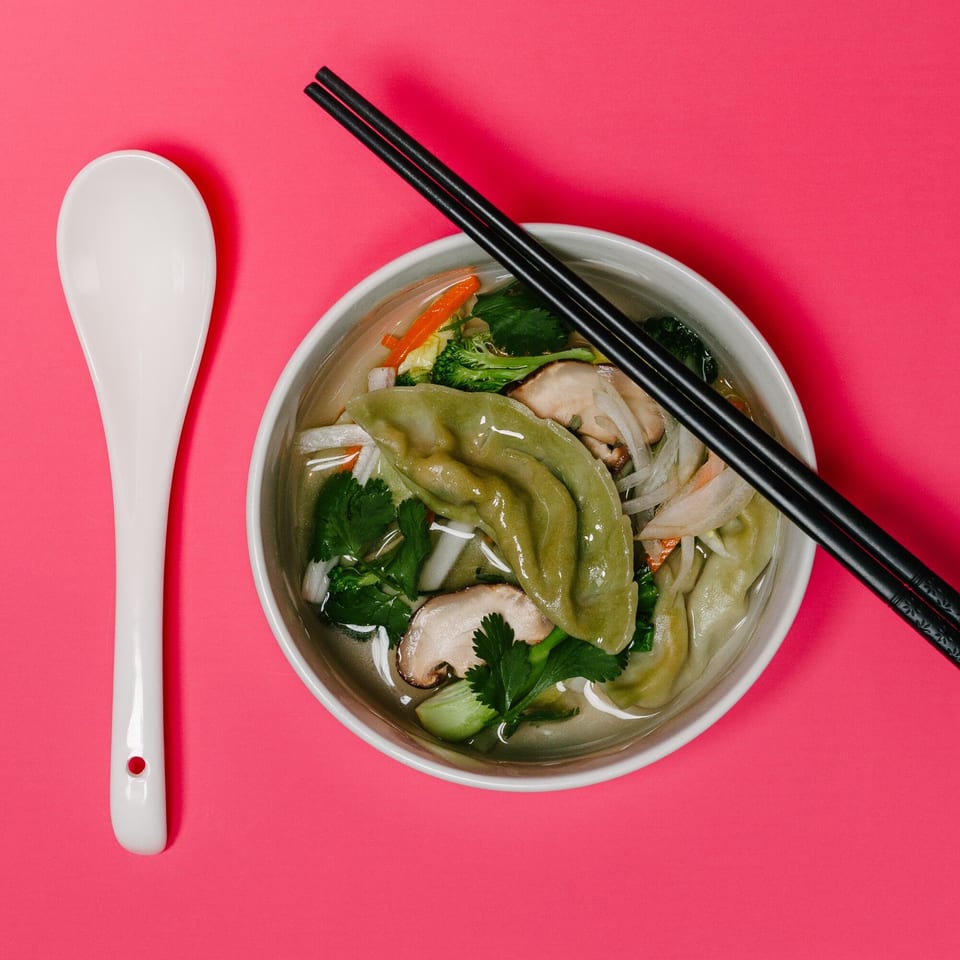 A bowl of Noods To Go dumpling soup with vegetables and chopsticks on a pink background.