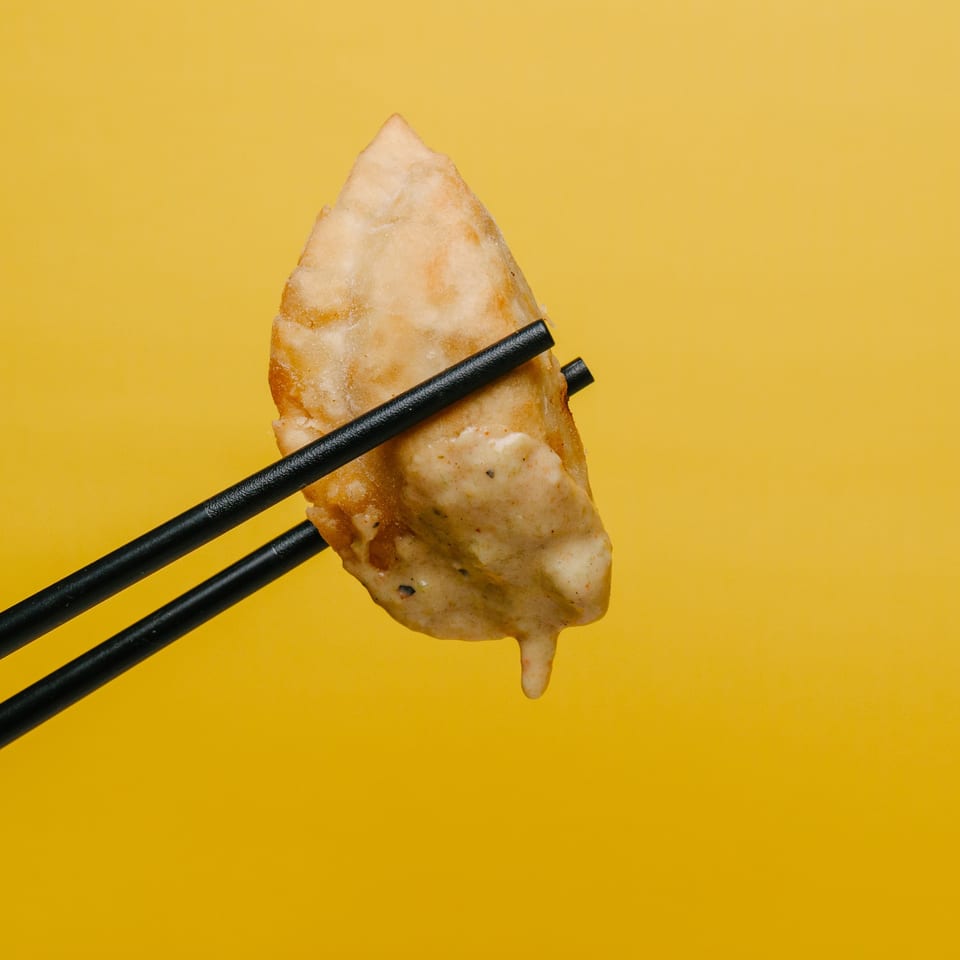 A fried dumpling with sauce dripping, held by black chopsticks against a yellow background.