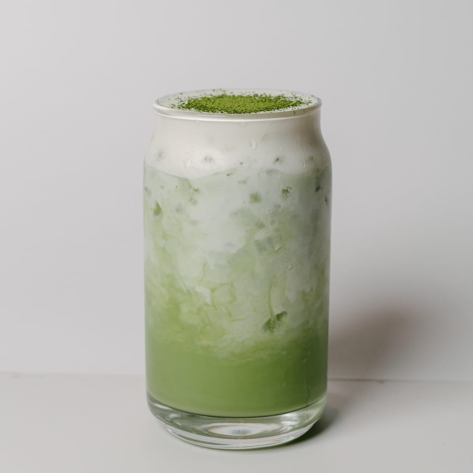 Iced matcha latte with a thick layer of foam and green powder on top, in a clear glass.
