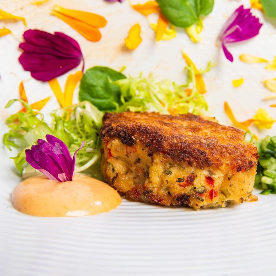 Crispy golden-brown crab cake with fresh greens, orange sauce, and edible flower petals on a white plate.