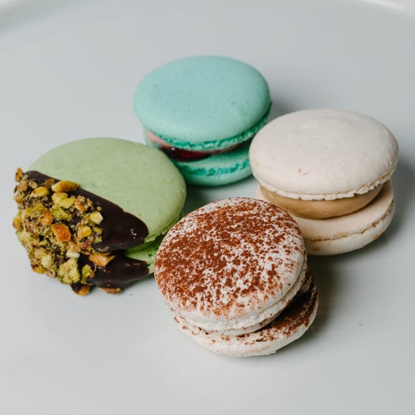 Four colorful macarons on a white plate, including pistachio, tiramisu, and blue raspberry flavors.