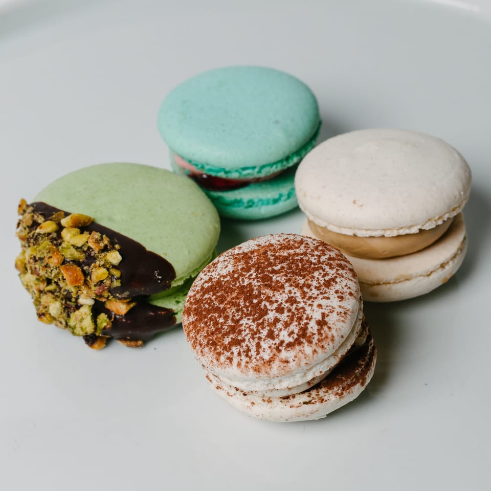 Four colorful macarons on a white plate, including pistachio, tiramisu, and blue raspberry flavors.
