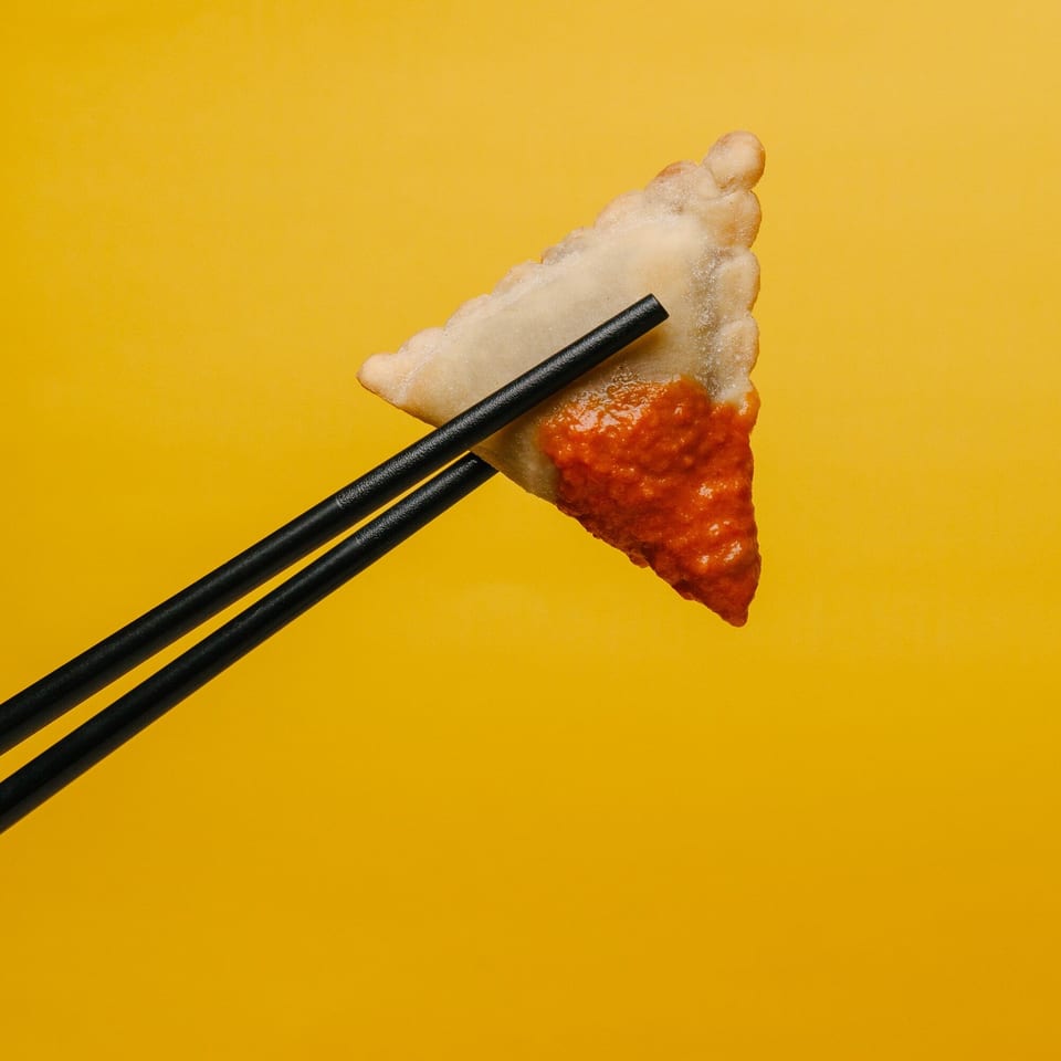 A triangular fried dumpling with red sauce, held by black chopsticks against a yellow background.