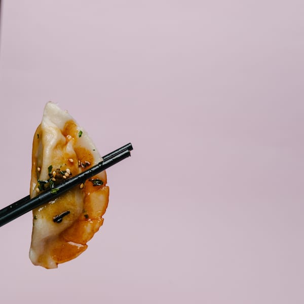 A single Noods To Go dumpling, glazed and topped with sesame seeds and herbs, held by black chopsticks.