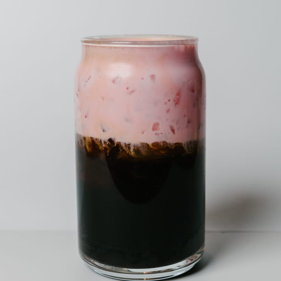 A glass of Noods To Go's dark iced coffee topped with a thick layer of pink foam.