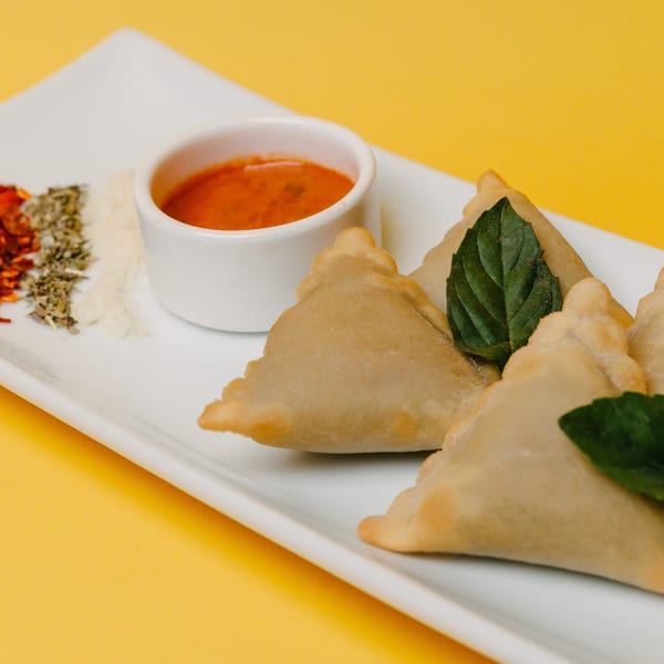 Golden-brown samosas with fresh basil, a side of red dipping sauce, and spices on a white plate.