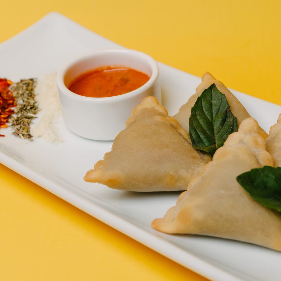 Golden-brown samosas with fresh basil, a side of red dipping sauce, and spices on a white plate.