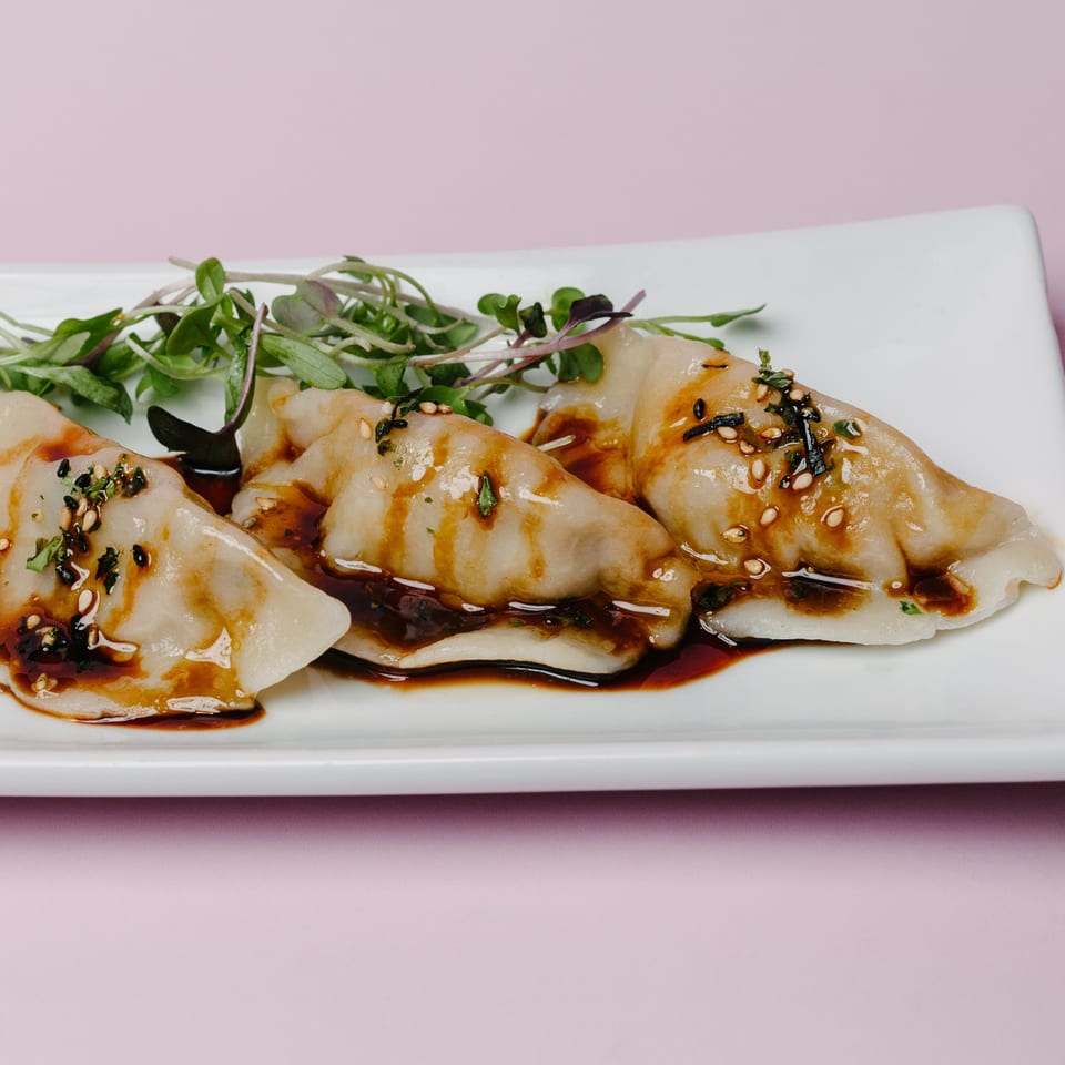 Three Asian-fusion dumplings drizzled with sauce and garnished with microgreens on a white plate.