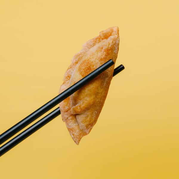 A golden-brown fried dumpling, held by black chopsticks, against a bright yellow background.