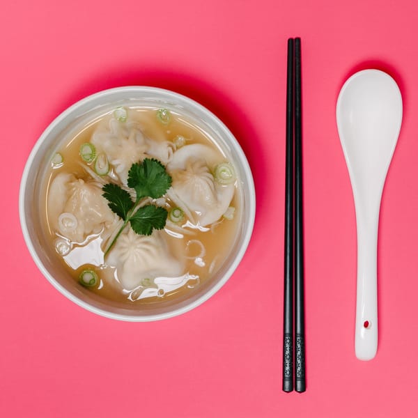 A bowl of Noods To Go wonton soup with cilantro and green onions, chopsticks, and a spoon.
