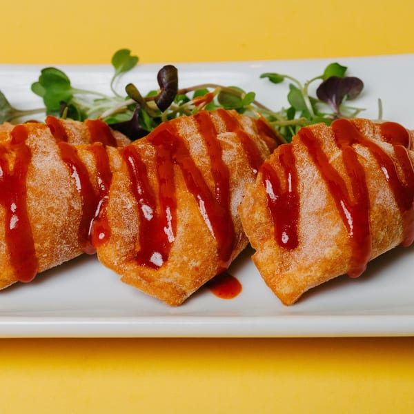 Four crispy Asian-fusion dumplings drizzled with red sauce and garnished with microgreens on a white plate.