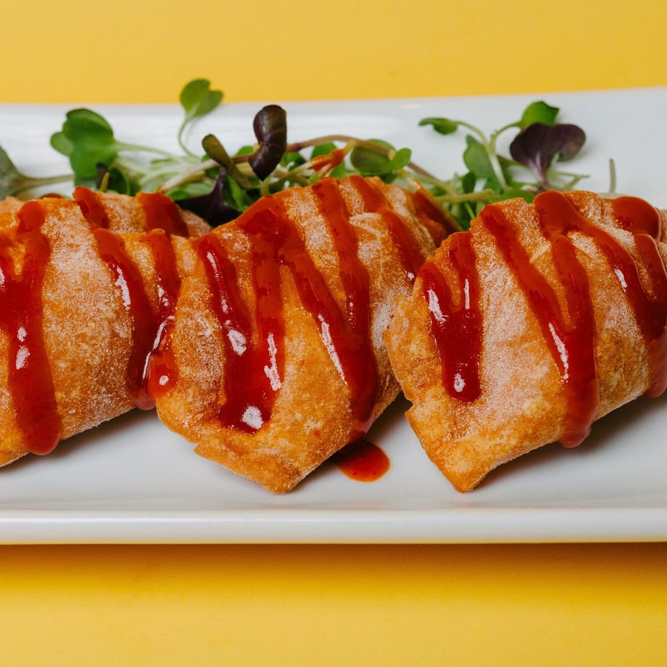 Four crispy Asian-fusion dumplings drizzled with red sauce and garnished with microgreens on a white plate.